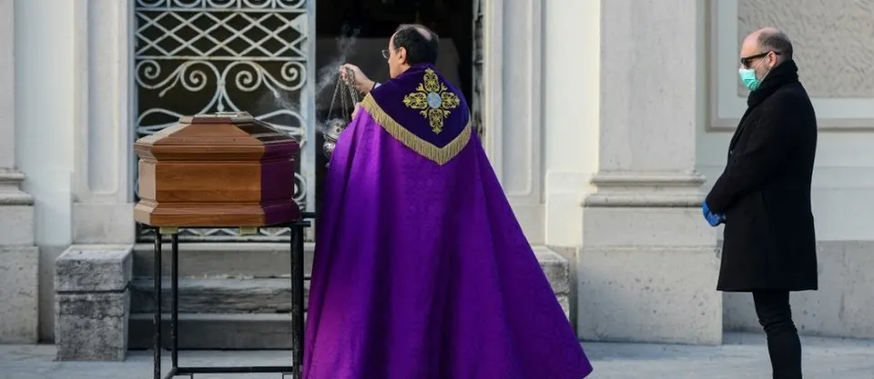 Rite en cours autours d'un cercueil par un homme religieux habillé de violet, avec une personne endeuillée regardant l'autre homme. Il porte un masque chirurgical et des gants pour se protéger du coronavirus