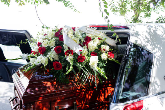 Cercueil sortant d'un coffre ouvert avec une couronnes de fleurs rouges et blanches et la mention 